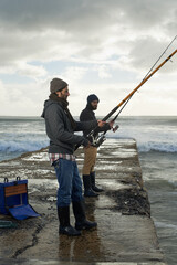 People, fishing and friends at beach on weekend, relaxing and casting a line by ocean. Men, fisherman and cloudy sky on vacation or holiday, hobby and bonding by wave and support on outdoor adventure