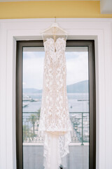 Lace wedding dress with straps hanging on a hanger on the balcony with the reflection of the sea and mountains in the glass