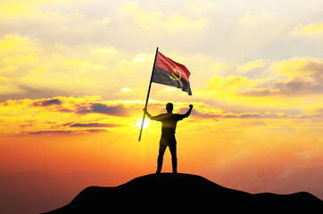 Angola flag being waved by a man celebrating success at the top of a mountain against sunset or sunrise. Angola flag for Independence Day.