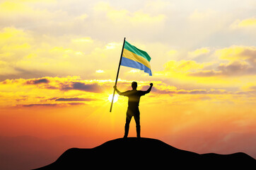 Gabon flag being waved by a man celebrating success at the top of a mountain against sunset or sunrise. Gabon flag for Independence Day.