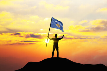 Kosovo flag being waved by a man celebrating success at the top of a mountain against sunset or sunrise. Kosovo flag for Independence Day.