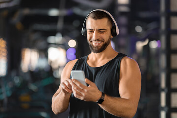 Handsome Muscular Male Athlete In Wireless Headphones Using Mobile Phone At Gym