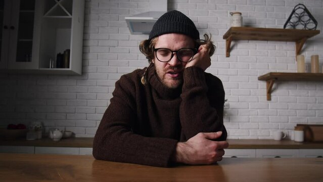 Tired young guy has no energy. Sleeping guy at dining room. Overworked man sleeping on folded hands in kitchen. Bored just fall asleep at home. Drunk slumber male in sweater and hat