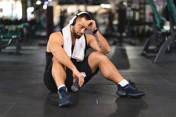 Fototapeta na wymiar Tired man sitting on gym floor with headphones on, relaxing after training