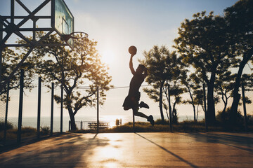 Naklejka premium black man doing sports, playing basketball on sunrise, jumping silhouette