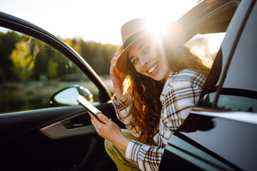 Young beautiful woman traveling by car using smartphone at sunset. Leisure, travel, technology,...