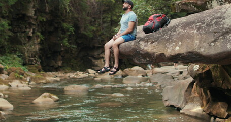 A lonely man-traveler calmly sits and rests on a stone that hangs over the water near the river against the background of the forest and the riverbed.