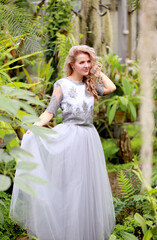 A blonde beauty poses in a gray tulle dress in a botanical garden. Background greenery and tropical plants. Beauty and fashion concept