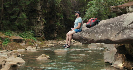 A lonely man-traveler calmly sits and rests on a stone that hangs over the water near the river against the background of the forest and the riverbed.