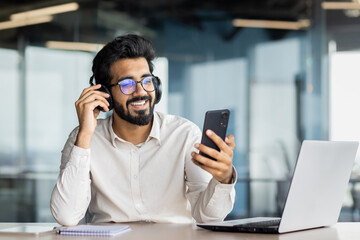 A smiling young Indian man is sitting in the office at a desk with a laptop, wearing headphones and using a mobile phone. Talks on a video call, listens to music during a work break