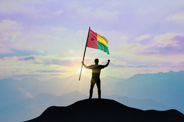 Guinea-Bissau flag being waved by a man celebrating success at the top of a mountain against sunset or sunrise. Guinea-Bissau flag for Independence Day.