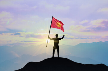 Kyrgyzstan flag being waved by a man celebrating success at the top of a mountain against sunset or sunrise. Kyrgyzstan flag for Independence Day.