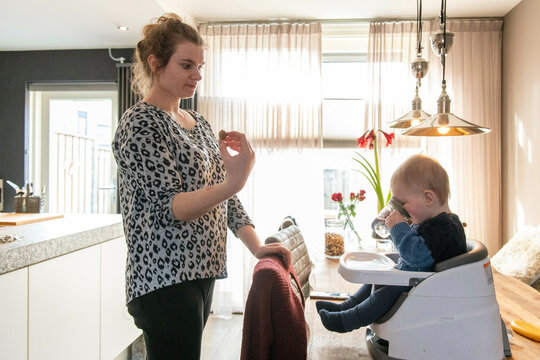 Mother feeding her small baby his lunch while he sits in his high chair on top of the kitchen side. Playing with the family dog
