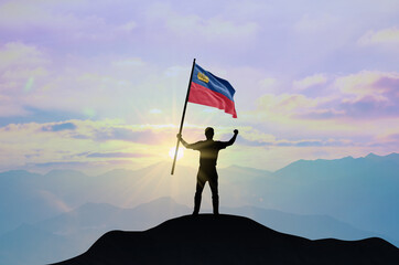 Liechtenstein flag being waved by a man celebrating success at the top of a mountain against sunset or sunrise. Liechtenstein flag for Independence Day.