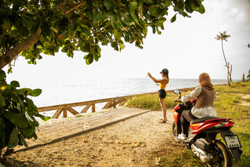 Two young female friends one wearing a hijab, stop their moped to take a photo of the coastal view