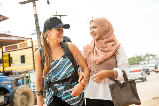 Two young women one wearing a Hijab walk together arm in arm smiling  in a rural town in Thailan