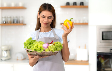 Young adult smiling lovely housewife standing in kitchen preparing fresh healthy vegan salad essential green vegetables and trying new recipe. Enjoyment dieting healthy nutrition lifestyle.