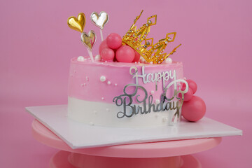 Pink birthday cake with gold princess crown and hearts on pink background close up.	