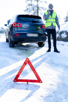 Close up of warning triangle on the road and driver wearing safety vest and calling roadside assistance while standing next to his car on winter day.