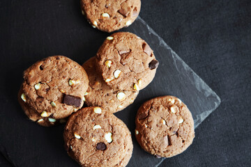 Chocolate cookies next to dark chocolate slices with nuts on a black stand on a dark background