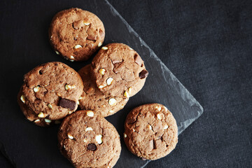 Chocolate cookies next to dark chocolate slices with nuts on a black stand on a dark background