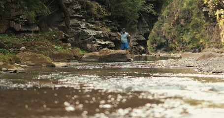 A man traveler drinks water sitting on a stone near a river in a summer forest. A male tourist with a backpack rests by the river during a hiking trip in the mountains.