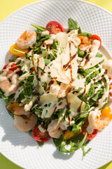 Fresh salad with greens, tomatoes, grilled shrimps on white plate. Top view on yellow background.