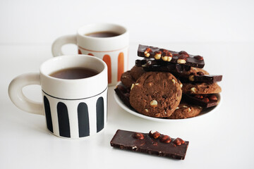 Chocolate cookies with chocolate slices and nuts on a plate next to two cups of tea on a white background