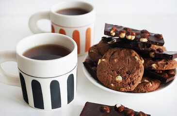 Chocolate cookies with chocolate slices and nuts on a plate next to two cups of tea on a white background