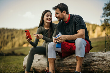 Diverse couple in fitness gear chatting happily while resting after a jog on a hill road, holding a smartphone