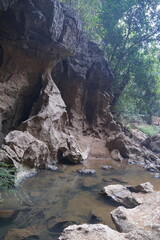 A large cave in the forest surrounded by large rocks and water flowing from the mouth of the cave.