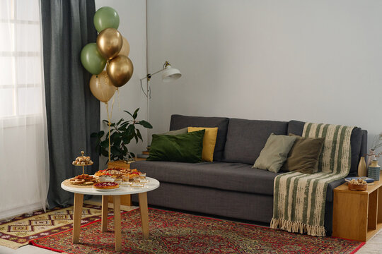 Corner of cozy living room with comfortable couch with cushions standing next to small table with snacks prepared for celebrating Eid al-Fitr