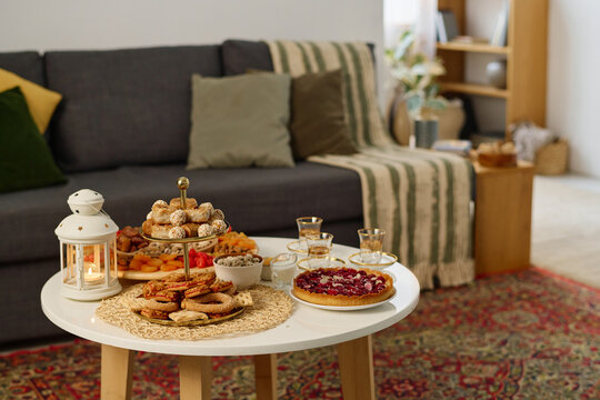 Traditional Eid al-Fitr baklava, cookies, sweet pie and other snacks standing on small round table against sofa with cushions in living room
