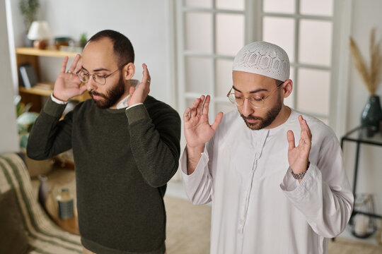 Young Muslim twin brothers keeping eyes closed and hands raised while standing in front of camera at home and saying namaz prayer