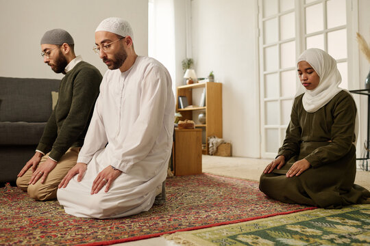 Young Muslim woman in hijab and two men standing on knees on carpet in living room and performing prayer on occasion of Eid al-Fitr