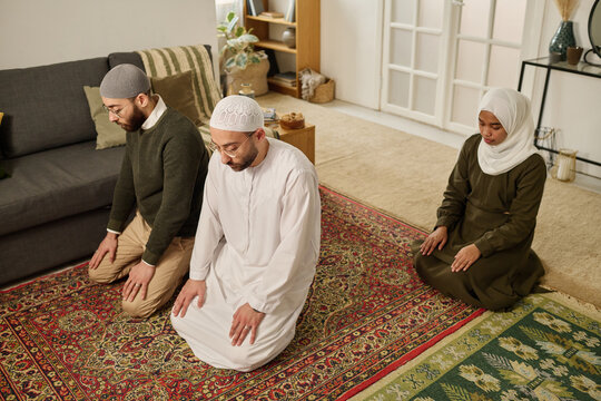 Two young religious men and woman in Muslim clothes standing on knees on the floor in living room and performing silent prayer