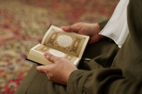 Hands of young unrecognizable woman in Muslim clothes holding open Holy Quran and reading verses of surah while sitting on the floor