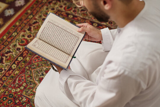 High angle of young Muslim man in thobe holding open Holy Quran with Arabic calligraphy and reading verses on religious occasion