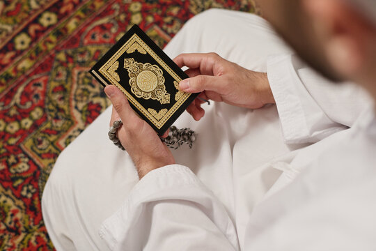 Hands of young Muslim man in white kandora standing on knees on the carpet and holding Holy Quran and rosary beads during silent pray