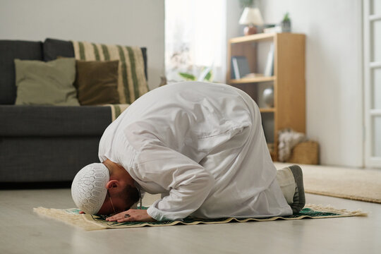Young Muslim man in skullcap and kandora bending forwards while standing on knees and touching rug by head during prayer