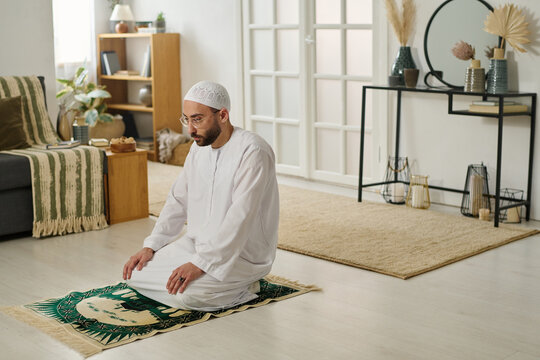 Young religious man in traditional Muslim skullcap and thobe standing on knees on rug in living room and performing silent prayer