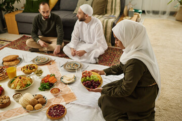 Muslim woman in white hijab standing on knees and putting plate with fresh grapes on tablecloth with national food prepared for dinner