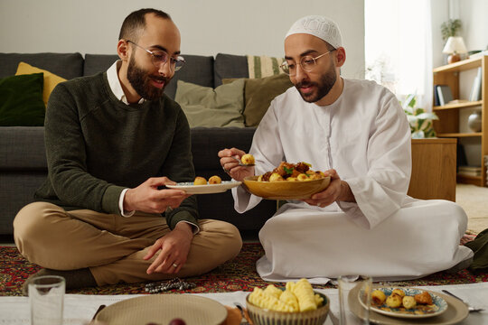 Young hospitable man in Muslim clothes holding plate with baked homemade meal and putting potato to his twin brother
