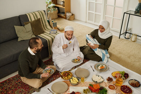 Two young Muslim men sitting on the floor in front of tablecloth with served homemade food, having baked potatoes and talking