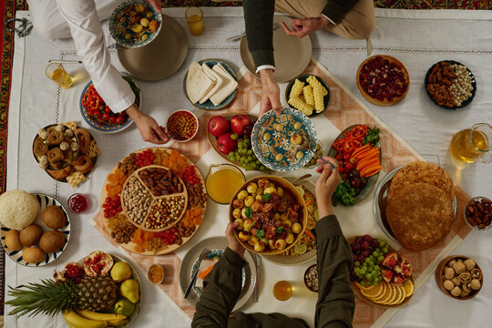 Top view of hands of young unrecognizable Muslim woman putting some baked potatoes on plate of guest over tablecloth with meals