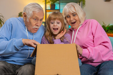Happy excited Caucasian grandparents and granddaughter girl opening cardboard box together sits on...