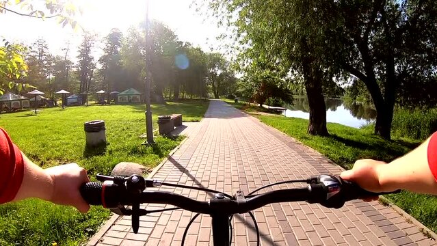 Persona woman riding bicycle along road made of paving slabs between green trees, bushes and grass on river bank on sunny summer day, brightly shining sun. POV. Man rides bicycle in park on river bank