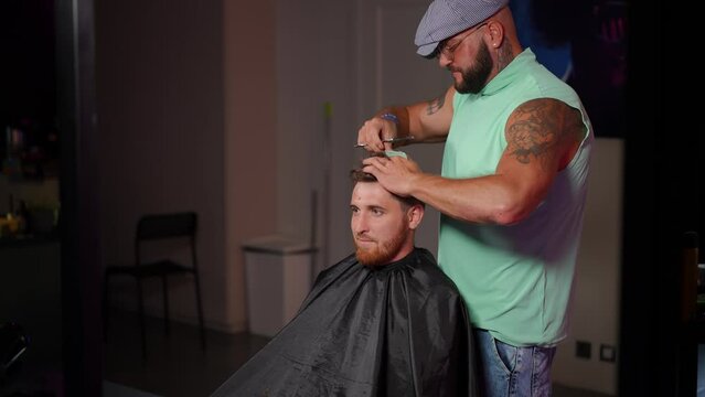 A stylish man in a cap cuts a haircut with barber scissors to a male client while standing in a training class. The process of learning hairdressing on real clients