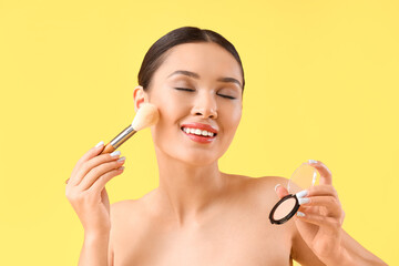 Obraz na płótnie Canvas Young Asian woman with makeup brush applying powder on yellow background, closeup