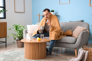 Ill young man with cup of tea and nasal drops sitting on sofa at home